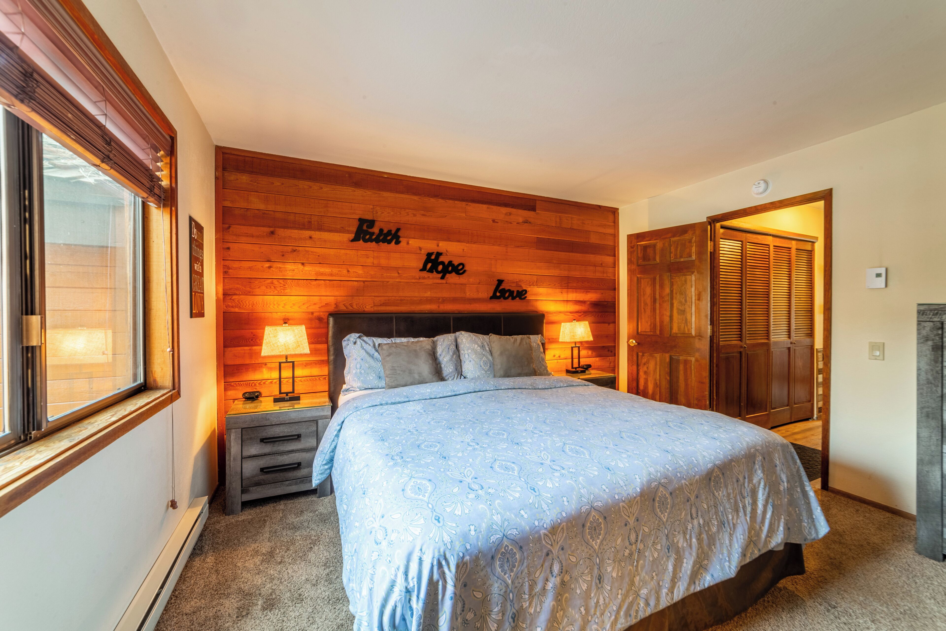 A bedroom with a wooden headboard, a bed with a patterned bedspread, two bedside lamps, and a window with blinds.