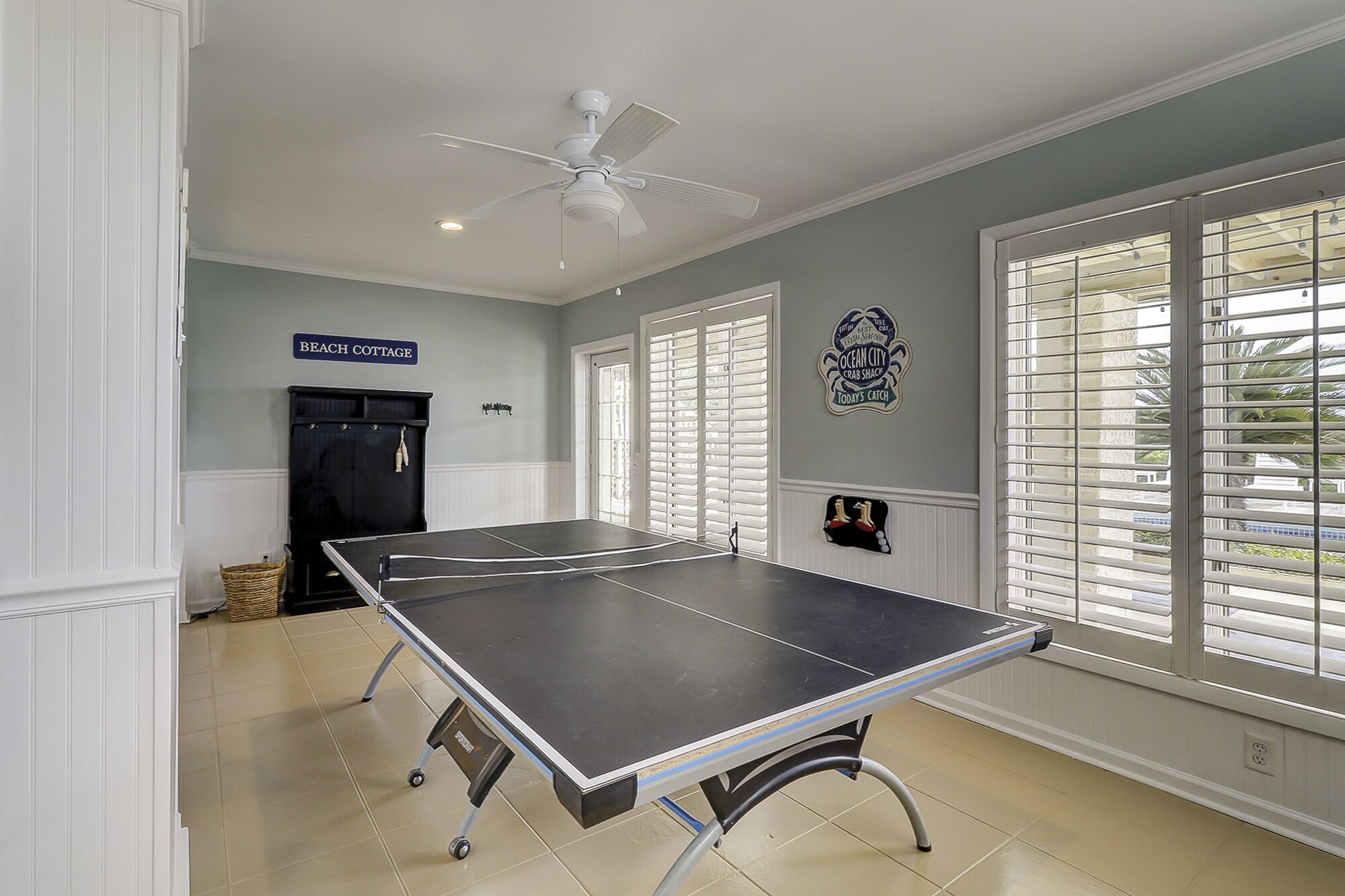 Une table de ping-pong dans une pièce dotée d’un ventilateur de plafond, une enseigne indiquant « CABANE DE PLAGE » et un élément décoratif mural.
