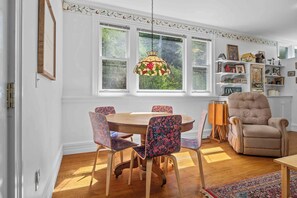 A dining area with a round wooden table, floral-patterned chairs, a hanging light fixture, and a bookshelf with various items. - Great Adventure: Family Fun Starts Here! (Keuka Park)