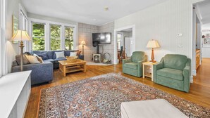 Een woonkamer met een blauwe bank, groene fauteuils, een houten salontafel, een kamerbreed tapijt en een flatscreen televisie aan de muur.