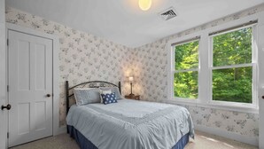 A bedroom with a bed, a nightstand, a lamp, and a window with a view of greenery.