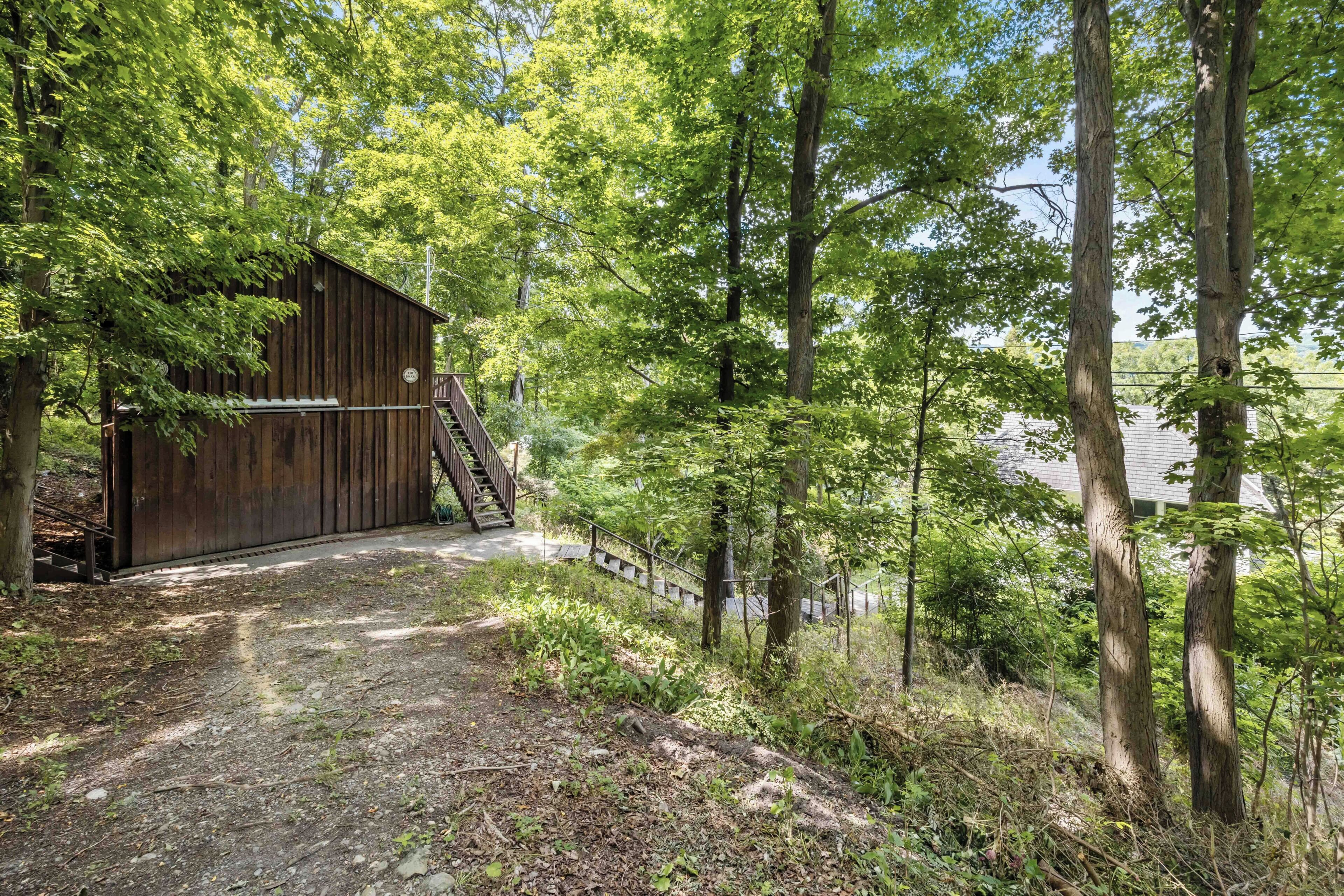 A rustic wooden barn with a wooden staircase leading up to a door, surrounded by dense green foliage and trees.