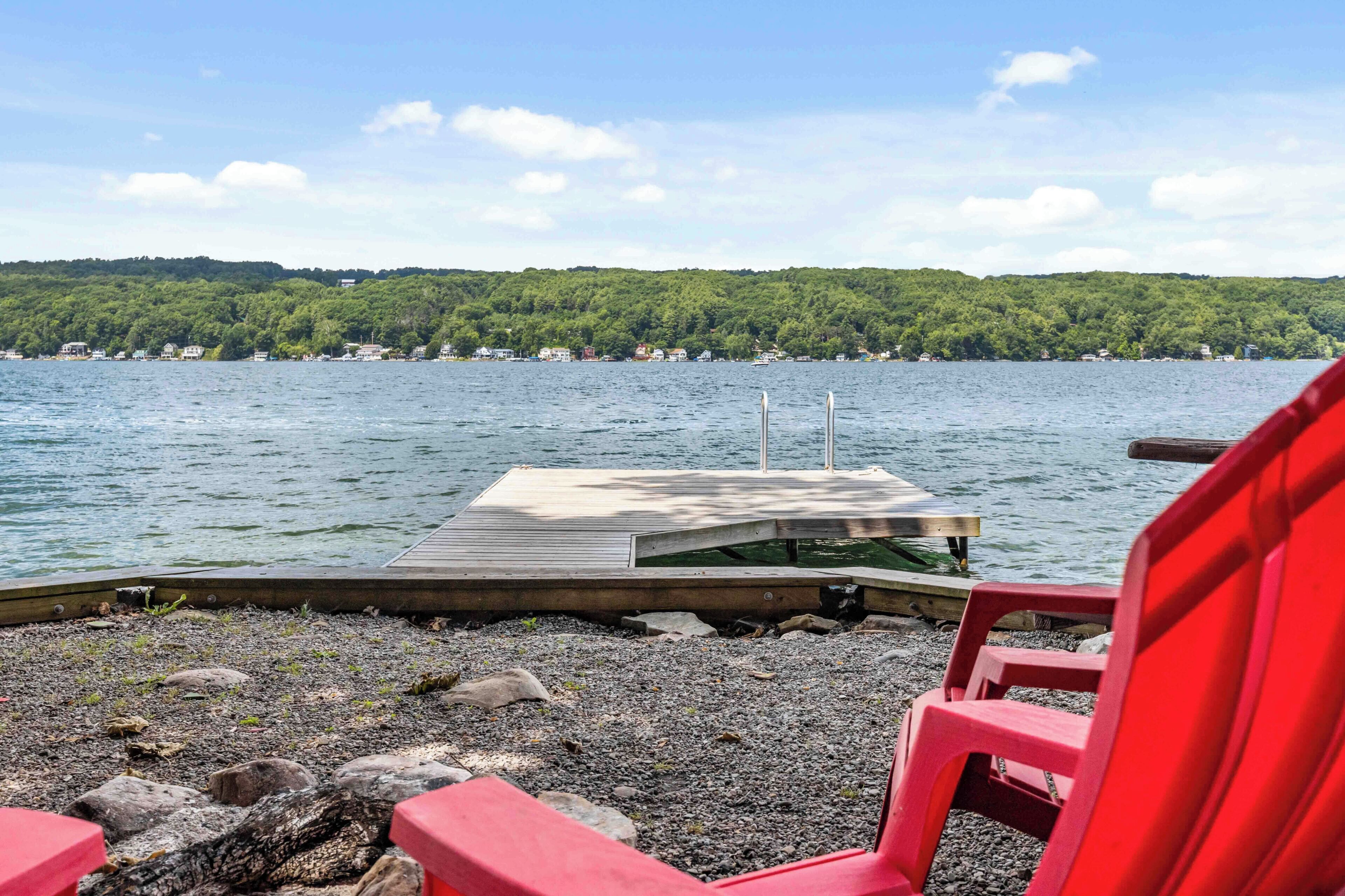 A lakeside view with a wooden dock extending into the water, surrounded by pebbles and a red chair.