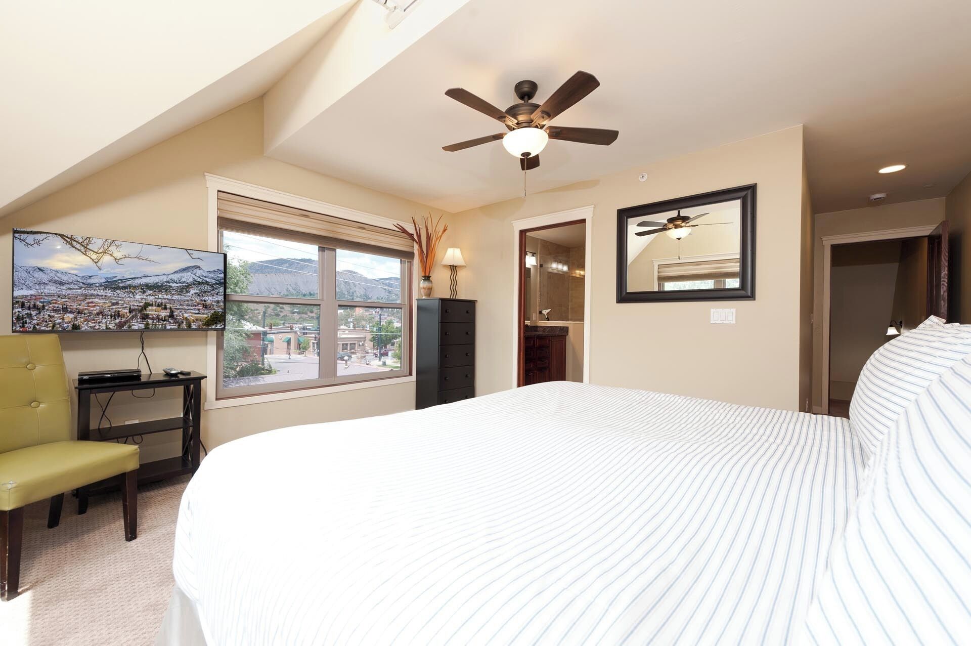 Une chambre à coucher comprenant un lit, un ventilateur de plafond, une télévision, une fenêtre avec vue et une commode.