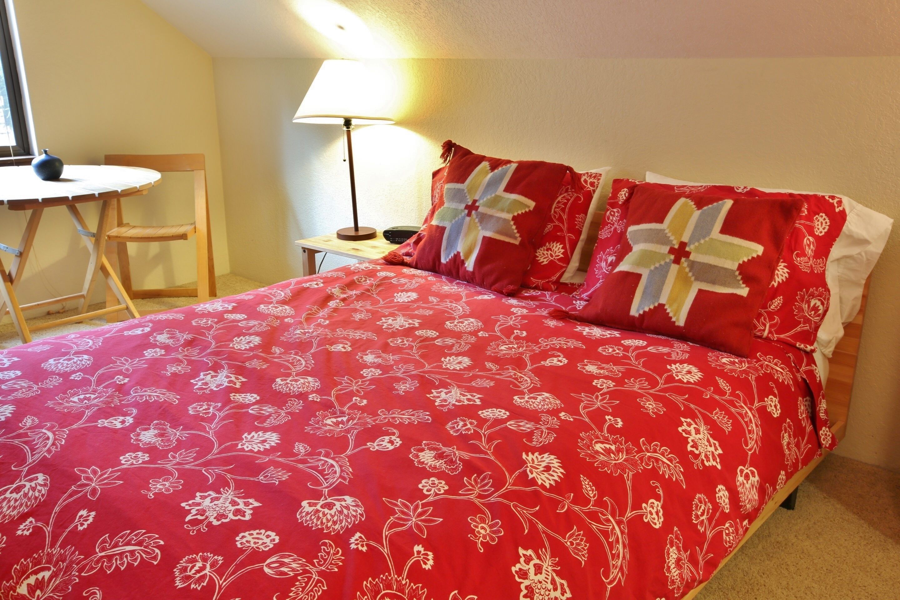A neatly made bed with a red floral bedspread, a small round table with a chair, and a bedside lamp.