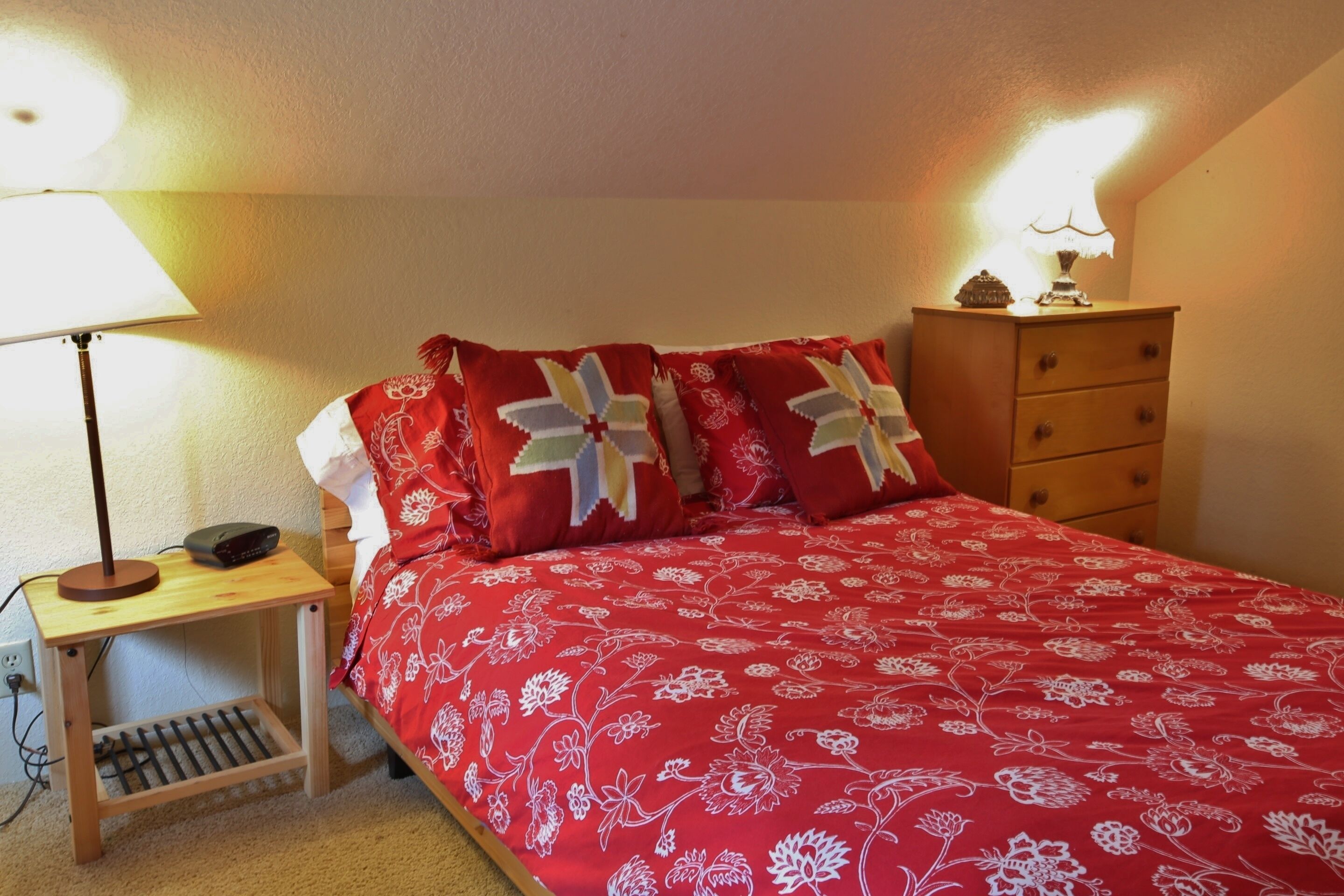 Um quarto com uma colcha vermelha florida, uma luminária de cabeceira, uma mesinha de cabeceira de madeira e um penteado com uma luminária em cima.
