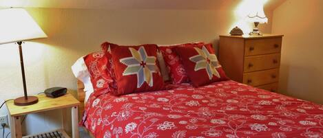 Um quarto com uma colcha vermelha florida, uma luminária de cabeceira, uma mesinha de cabeceira de madeira e um penteado com uma luminária em cima.