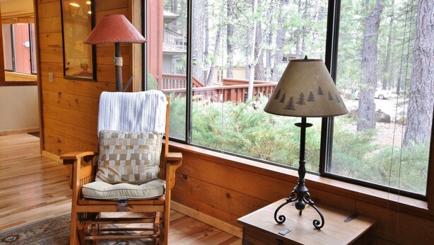 A wooden interior with a rocking chair, a table, and a lamp. A large window offers a view of a forested area. - 8 Leisure - Light & Bright Home, Hot Tub, Quiet Area Close to SHARC + Passes Included (Sunriver)