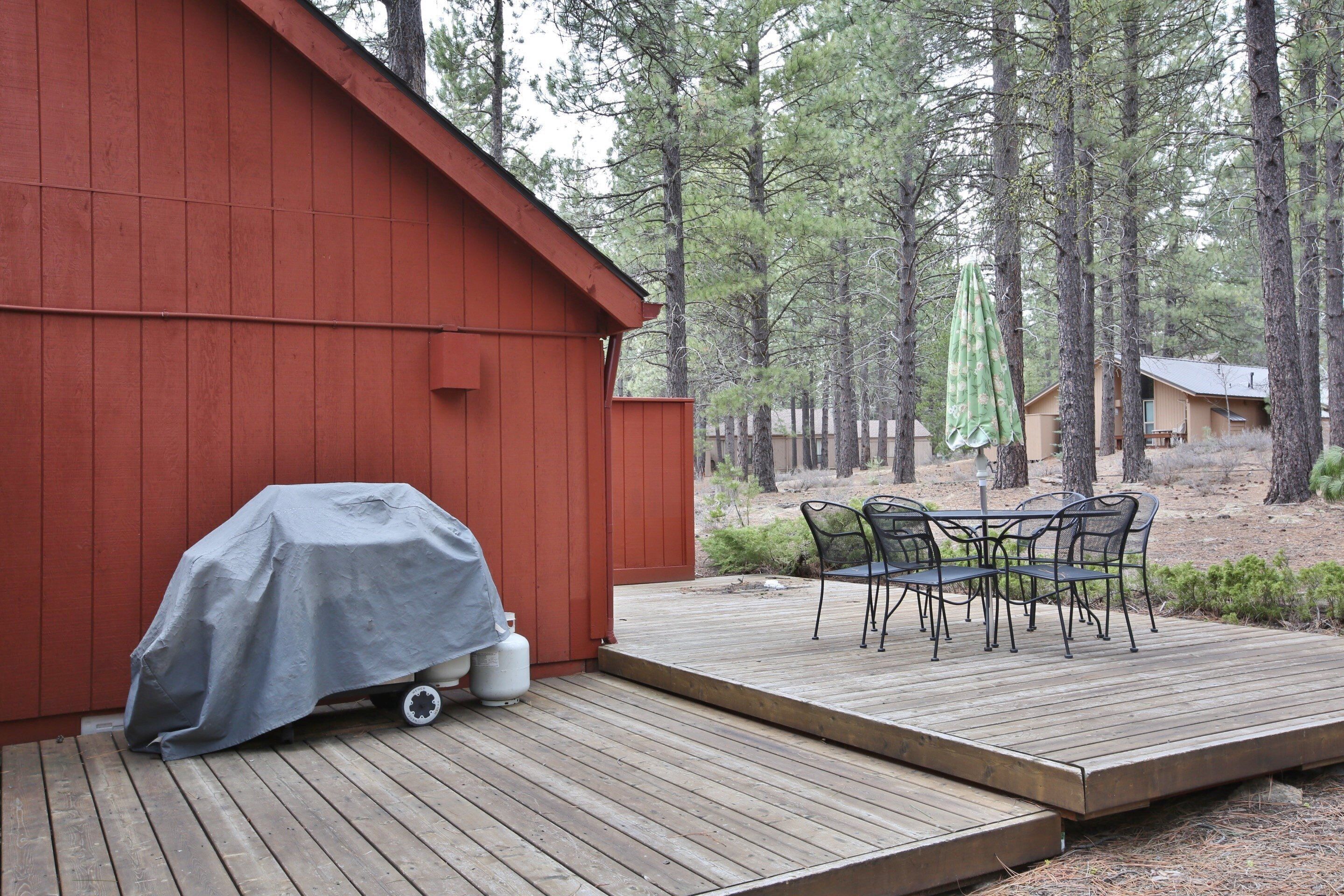 A wooden deck with a covered grill, a table with chairs, and a red building in a forested area.