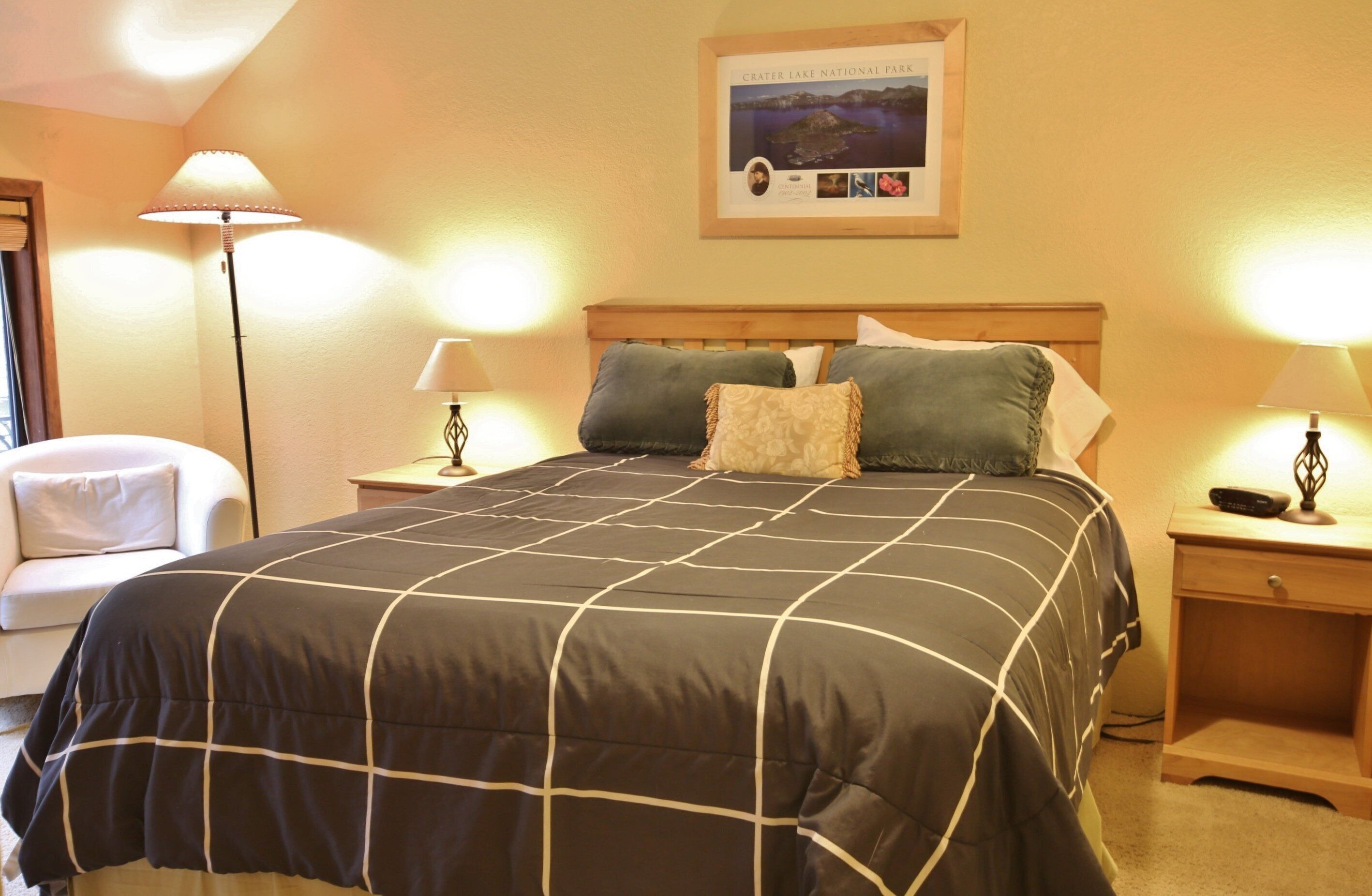 A neatly made bed with a black and white checkered bedspread, two pillows, and a framed picture of Crater Lake National Park on the wall.