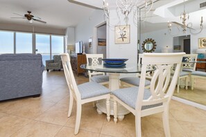 Un coin repas avec une table en verre entourée de chaises blanches dotées de coussins gris, un ventilateur de plafond et une grande fenêtre offrant une vue sur le ciel.