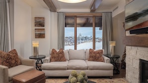 A cozy living room with a sofa, two side tables, a fireplace, and a large window with a snowy view. - Ultra Luxury + Pool + Slopeside - Snowline Ridge (Steamboat Springs)