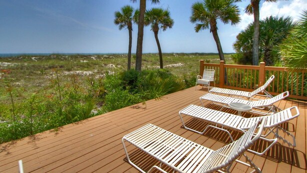 A wooden deck with white metal sun loungers and a wooden railing overlooking a beach with palm trees. - 2315 Windsor II -Beautiful Oceanfront 1 Bedroom Villa! (Hilton Head Island)