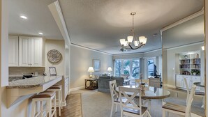 A spacious living area with a dining table, chairs, and a chandelier. There is a kitchen with white cabinets and a granite countertop. - 2315 Windsor II -Beautiful Oceanfront 1 Bedroom Villa! (Hilton Head Island)