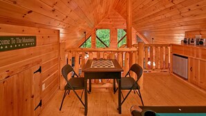 Interior de una cabaña de madera con una mesa de ajedrez, dos sillas, un letrero que dice "Welcome to The Mountains" y una zona de cocina con un refrigerador.