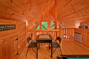 A wooden cabin interior with a chess table, two chairs, a sign reading "Welcome to The Mountains", and a kitchen area with a refrigerator.