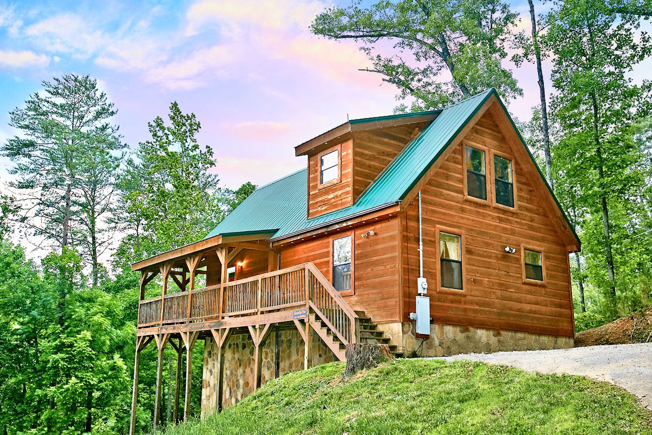A wooden cabin with a balcony, a stone foundation, and a green roof, surrounded by trees.