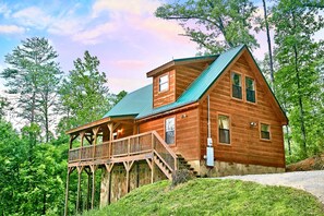 A wooden cabin with a balcony, a stone foundation, and a green roof, surrounded by trees.