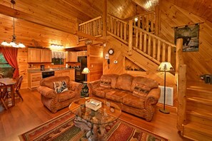 A log cabin interior with a dining area, two sofas, a coffee table, a kitchen, and a staircase.
