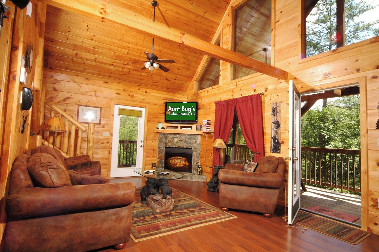 A cabin interior with a fireplace, two leather sofas, a ceiling fan, and a sign for 'Aunt Bug's Cabin Rentals, LLC.'