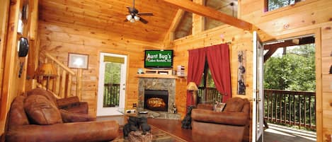 Interior de una cabaña con chimenea, dos sofás de cuero, un ventilador de techo y un letrero que dice 'Aunt Bug's Cabin Rentals, LLC'.