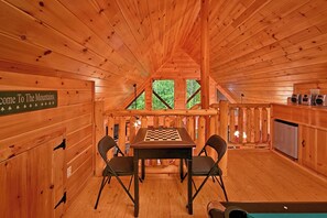 Interior de una cabaña de madera con una mesa de ajedrez, dos sillas, un letrero que dice "Welcome to The Mountains" y una zona de cocina con un refrigerador.