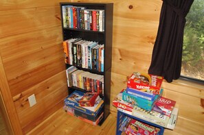 Una habitación con una estantería de madera repleta de libros, una ventana con cortina negra y una mesa con diversos juegos de mesa.