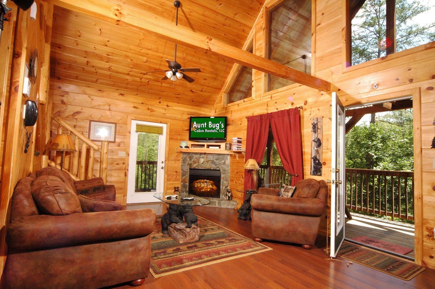 A cabin interior with a fireplace, two leather sofas, a ceiling fan, and a sign for 'Aunt Bug's Cabin Rentals, LLC.'