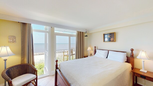 A hotel room with a large bed, two bedside lamps, a wicker chair, a view of the sea, and a framed picture on the wall.