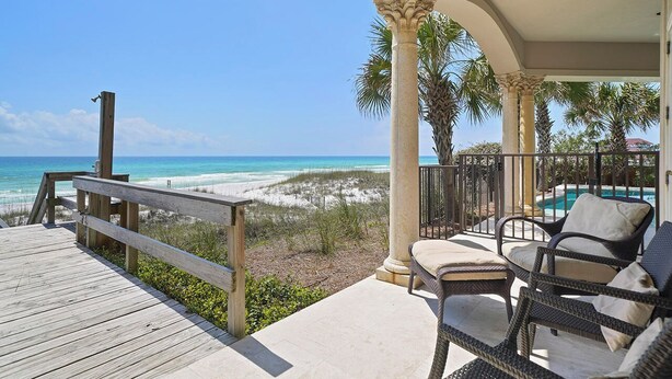 Une terrasse en bois offrant une vue sur la plage et l’océan, un patio couvert aménagé avec des meubles en osier et des palmiers.