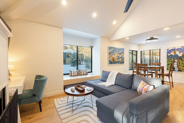 A modern living room with a grey sofa, wooden dining table, and chairs, a window with a view of trees, and artwork on the walls.
