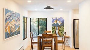 Uma sala de jantar com mesa e cadeiras de madeira, duas pinturas grandes nas paredes, uma janela com vista para árvores e uma geladeira de aço inoxidável.