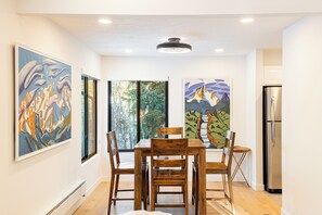 Un comedor con mesa y sillas de madera, dos pinturas grandes en las paredes, un ventanal con vista a árboles y un refrigerador de acero inoxidable.