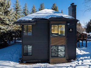 A small, single-story cabin with a snow-covered roof, large windows, and a chimney.