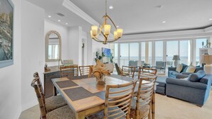 Un vaste espace dédié à la salle à manger, avec une table et des chaises en bois, un lustre et une vue sur l’océan grâce à de grandes fenêtres.
