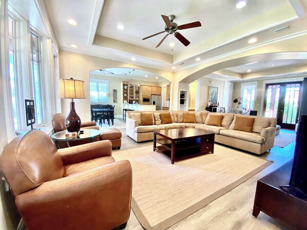 A spacious living room with a large sectional sofa, a coffee table, and a leather armchair. There is a ceiling fan, recessed lighting, and a view into the kitchen area.