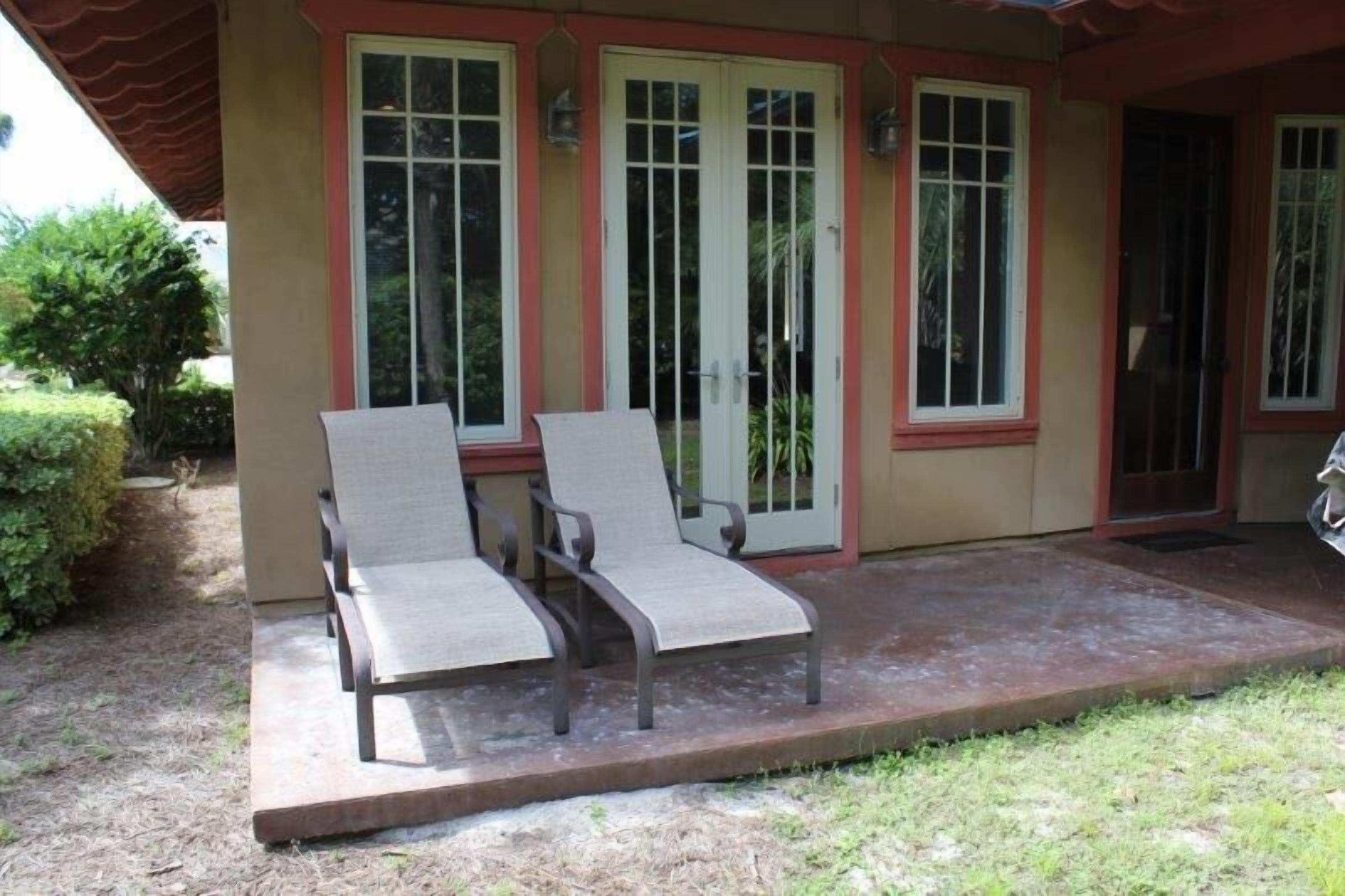 Un patio techado con dos sillones de descanso y una mesa pequeña, junto a un edificio con fachada roja y color crema y ventanas amplias.