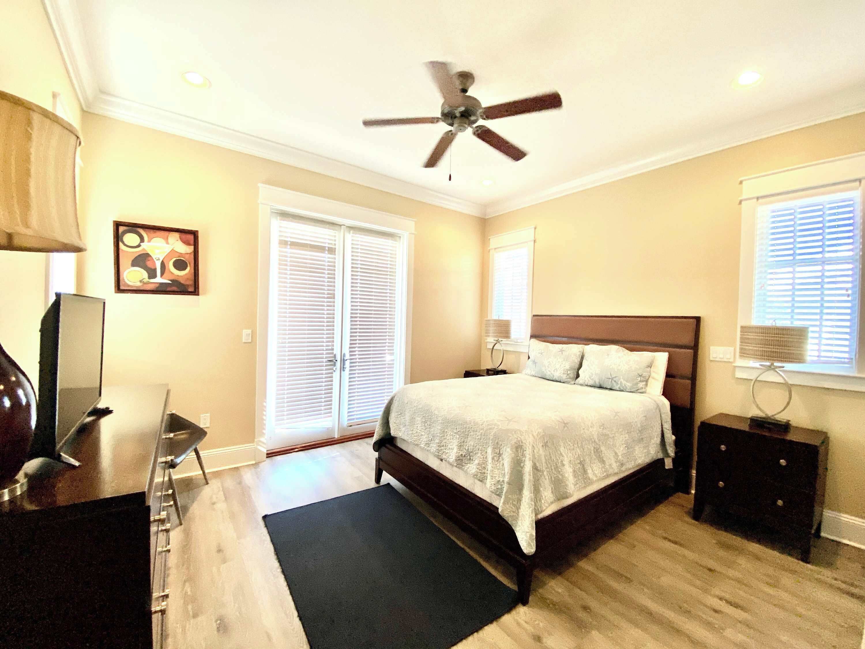 A bedroom with a bed, a ceiling fan, a desk, a chair, a TV, and a window with blinds.