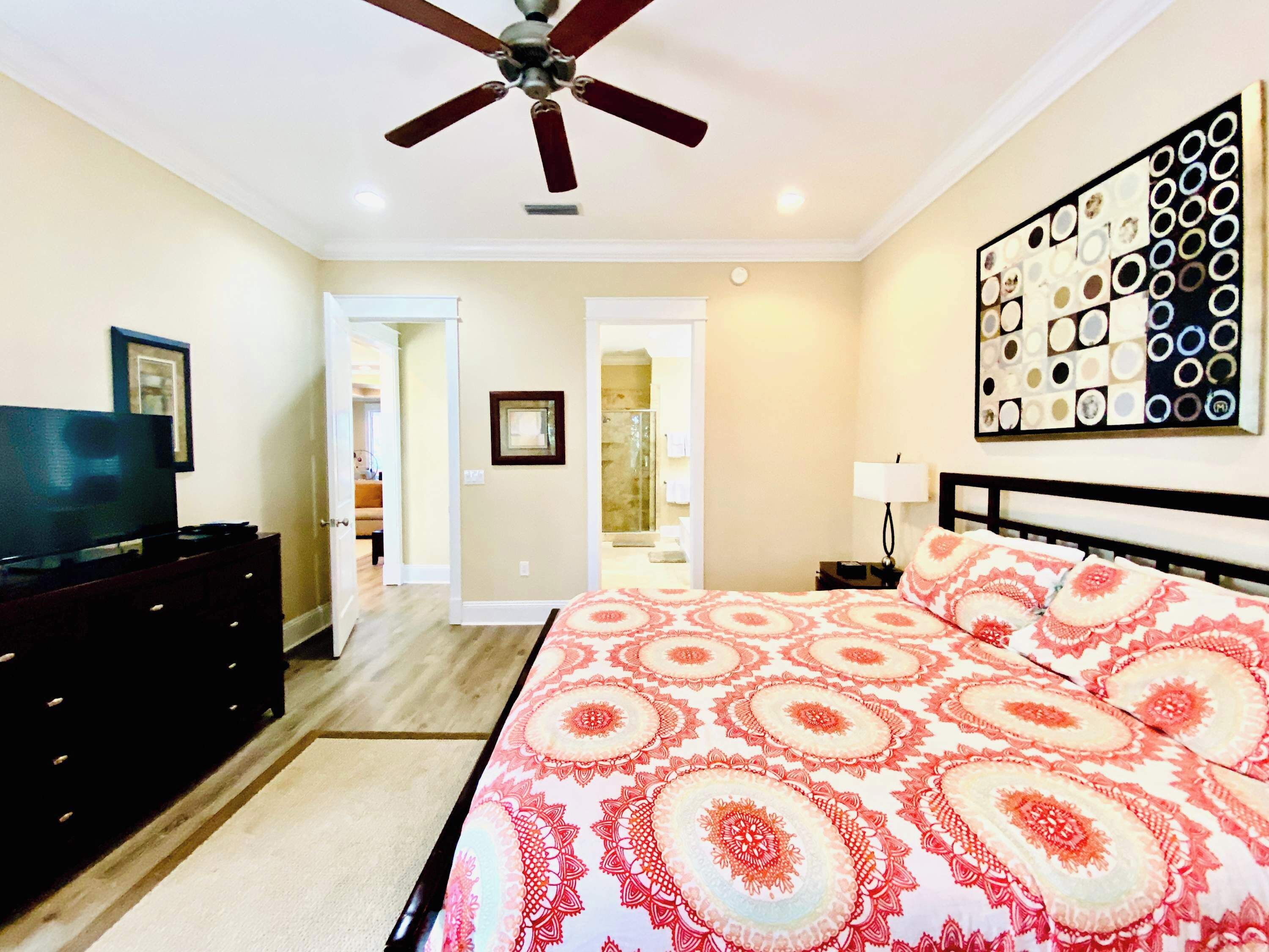 A bedroom with a bed, a ceiling fan, a TV on a dresser, and a hallway leading to a bathroom.