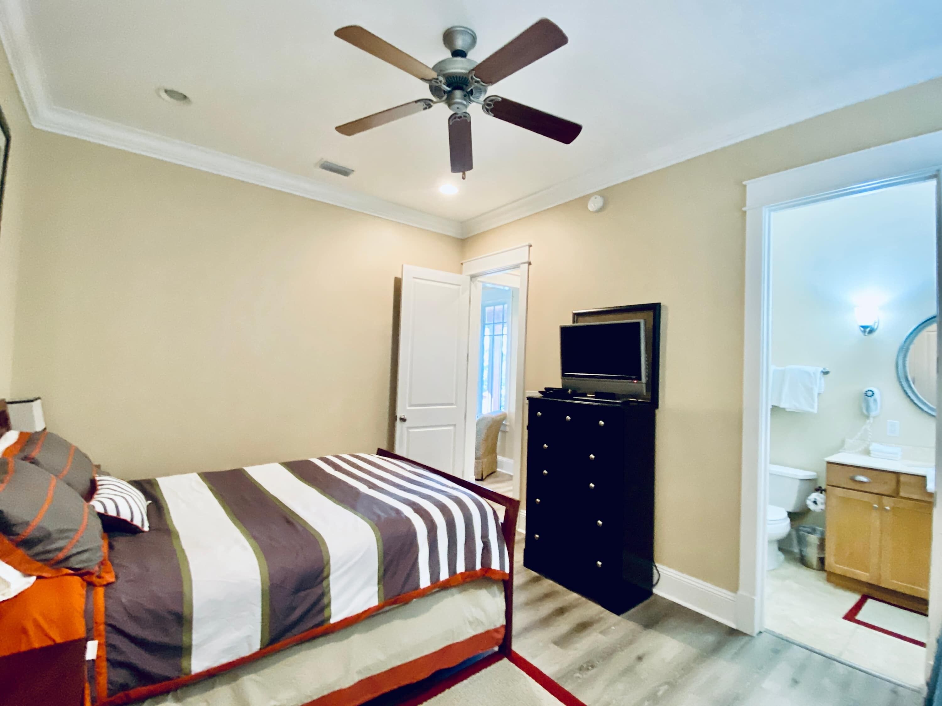A bedroom with a bed, a ceiling fan, a TV on a dresser, and a bathroom visible through an open door.