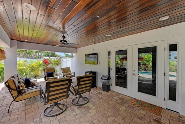 A covered patio with a wooden ceiling, outdoor seating, and a view of a pool.