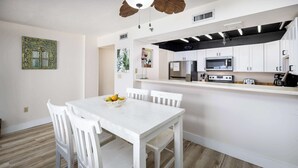 Een moderne keuken met witte kastjes, een magnetron en een koelkast. Er is een eettafel met witte stoelen en een schaal fruit.