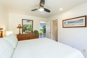 Una camera da letto con un letto, una ventola a soffitto, un comodino, una lampada, uno specchio, una finestra con vista, una porta e un quadro appeso al muro.