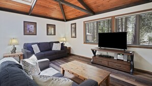 Un salon avec un plafond en bois, un canapé, une table basse, un téléviseur posé sur un meuble et une fenêtre avec des stores.