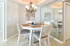 A dining area with a round table, white chairs, a chandelier, and artwork on the walls. - Tropics themed beachfront getaway w/private balcony! 🌴 (Destin)