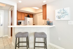 A kitchen with wooden cabinets, a white countertop, and two bar stools. - Tropics themed beachfront getaway w/private balcony! 🌴 (Destin)
