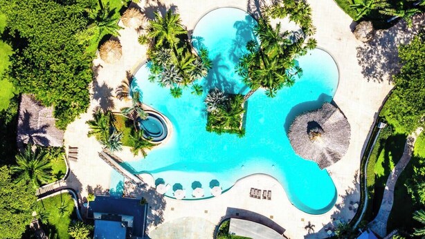 Vue aérienne d’un complexe hôtelier doté d’une grande piscine centrale, entourée de palmiers et de zones ombragées.