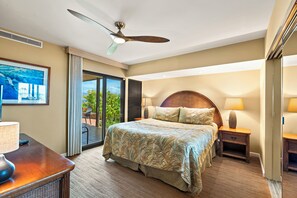 A hotel room with a bed, a desk with a lamp, a ceiling fan, and a view of the outdoors. - Wailea Elua 2003 - Ground Level Gem -Oceanfront Property W/ Tropical Grounds (Wailea, Maui)