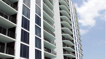 Un hotel moderno de gran altura con varios balcones, una zona de piscina con tumbonas y cielo despejado.