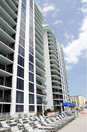 A modern hotel with multiple balconies, a pool area with sun loungers, and a clear sky. - Mermaid's Hideaway PCB-Beachfront-Family Friendly Vacation Rental (Panama City Beach)